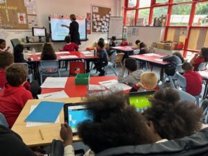 Lesson In the classroom at Mount Stuart Primary School. A teacher is stood at the front of the classrom surrounded by pupils at their desks.